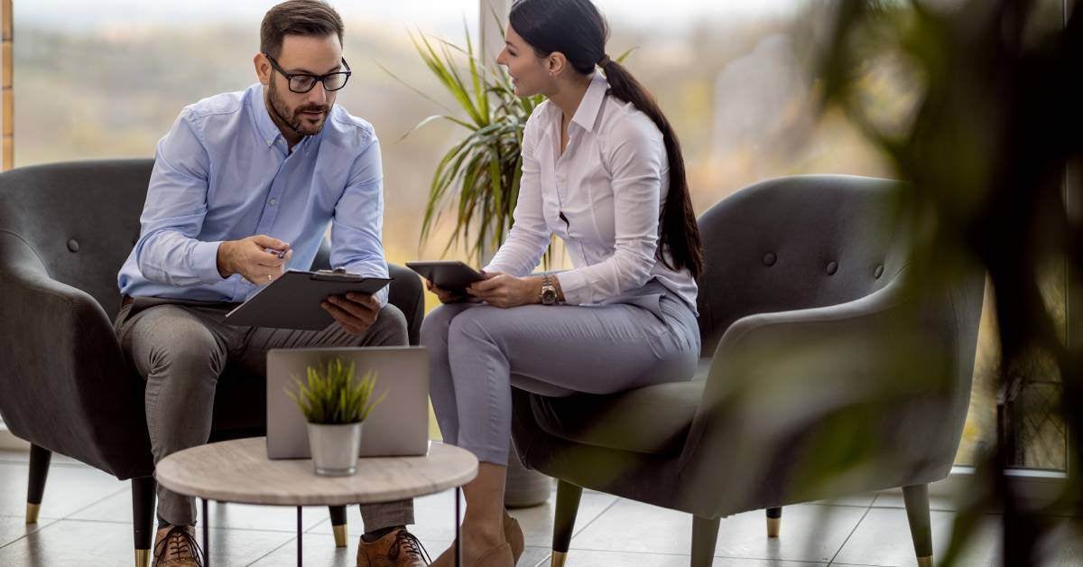 A young man and professional woman are sitting in chairs and going over information on a clipboard while closing a deal.