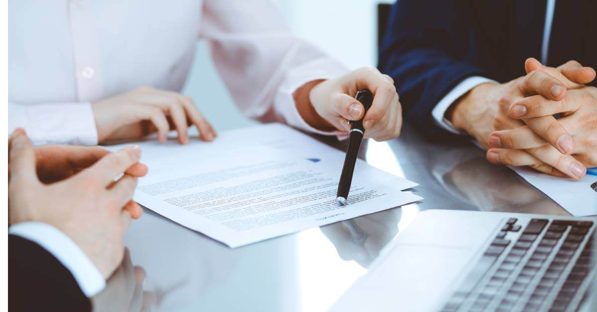 Three pairs of hands are visible on a table top, and a laptop is nearby. One hand points at a contract with a pen.