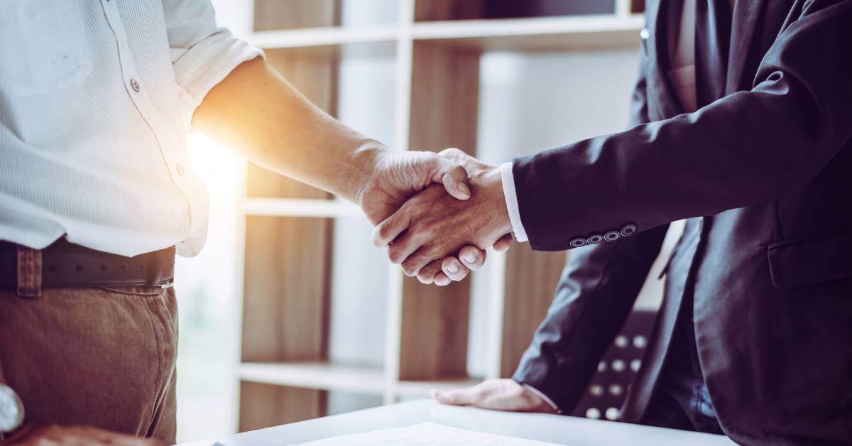 Two men in business attire are visible from the waist down. They are shaking hands over a newly signed contract.
