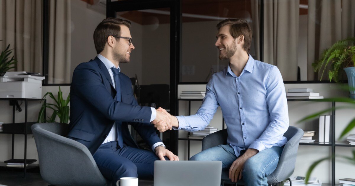Two young businessmen are sitting on chairs together. They are shaking hands to symbolize closing a business deal.