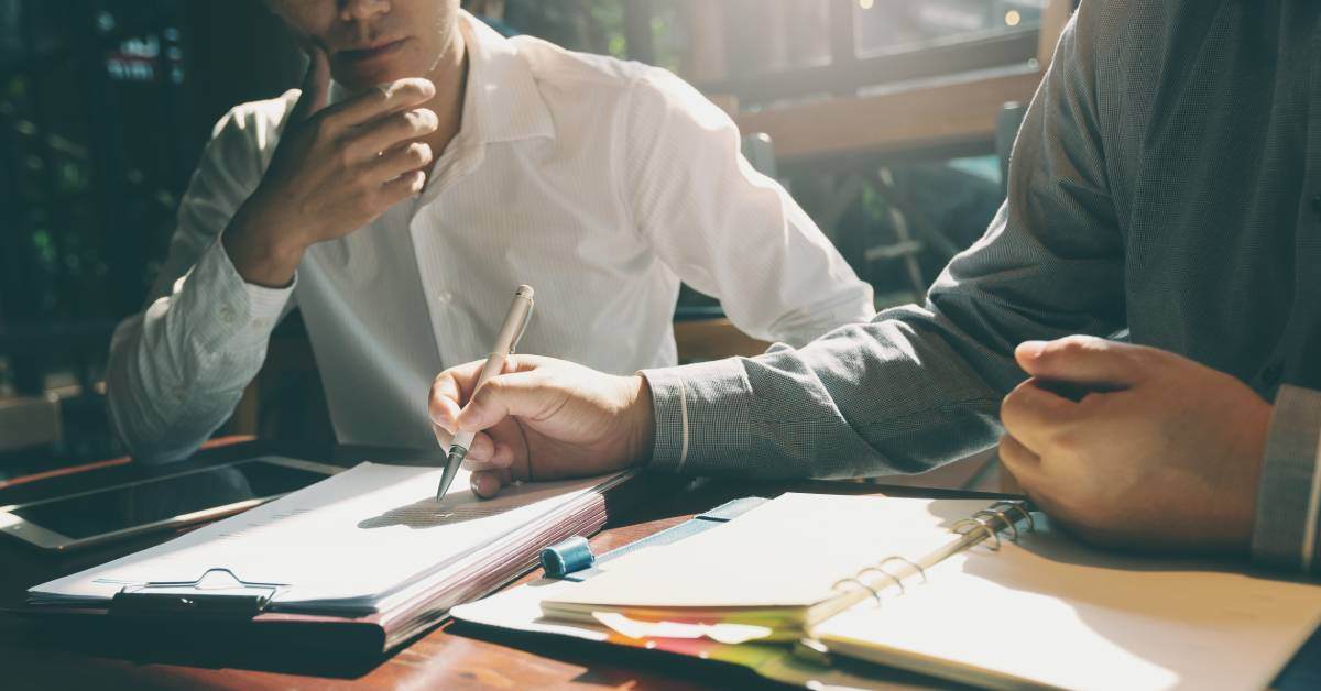 Two businessmen sitting at a desk and carefully going over several forms and folders with eyes and a pen.