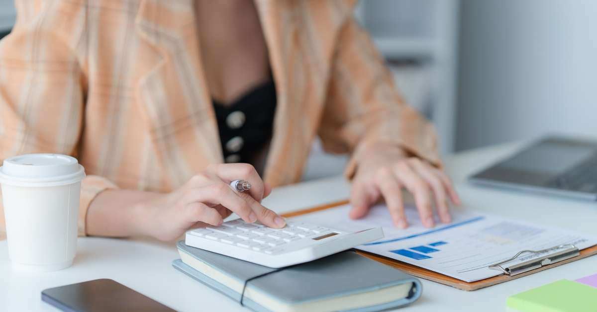 A woman in an orange blouse uses a calculator at a desk beside a clipboard with forms, a phone, and a coffee cup.