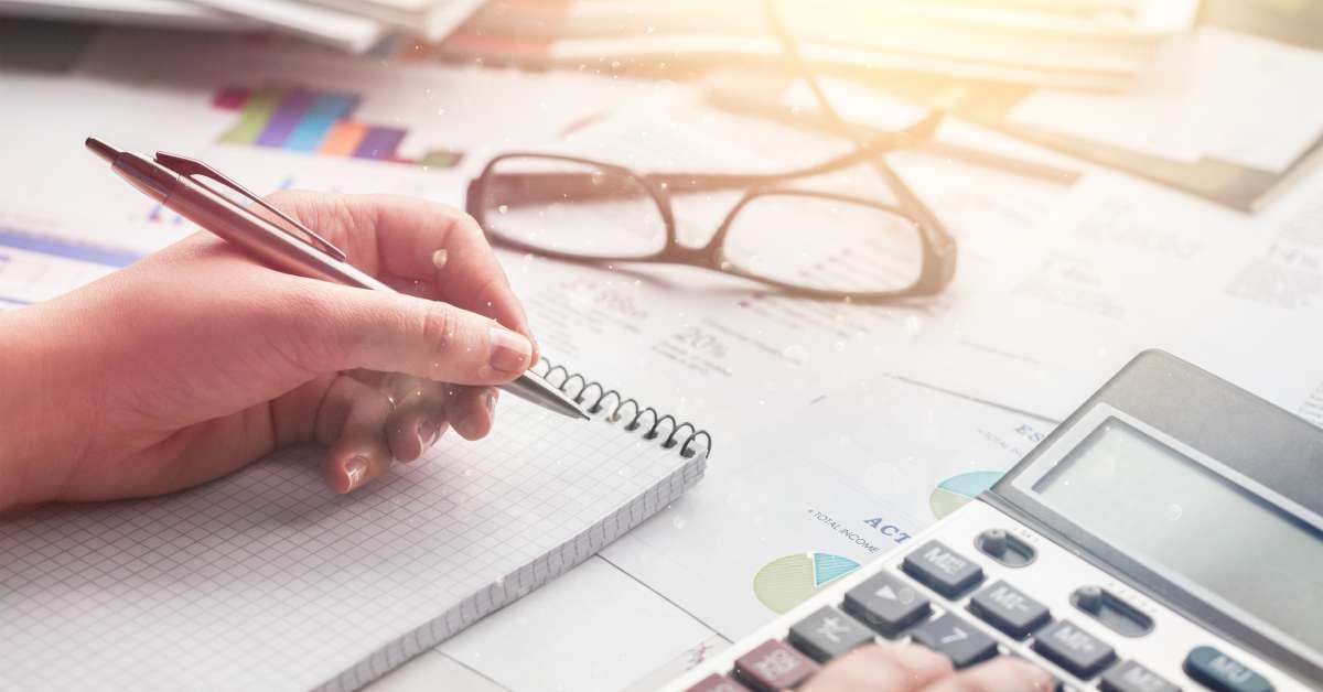 A desk with a calculator, a notepad, and glasses at close range as a person holds a pen and types on the calculator.