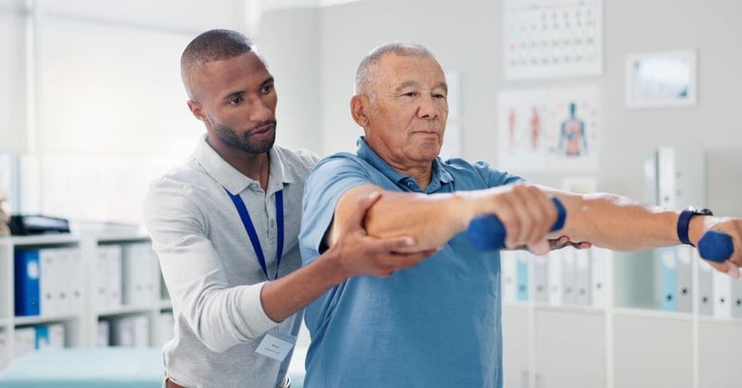 A young physical therapist assisting an older man with his therapy. The older man holds two small dumbbells in each hand.