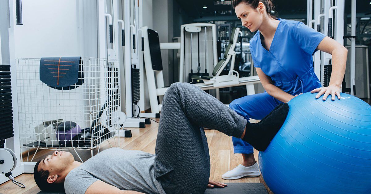 A young woman helping a man with his physical therapy using a blue exercise ball to work out his legs.