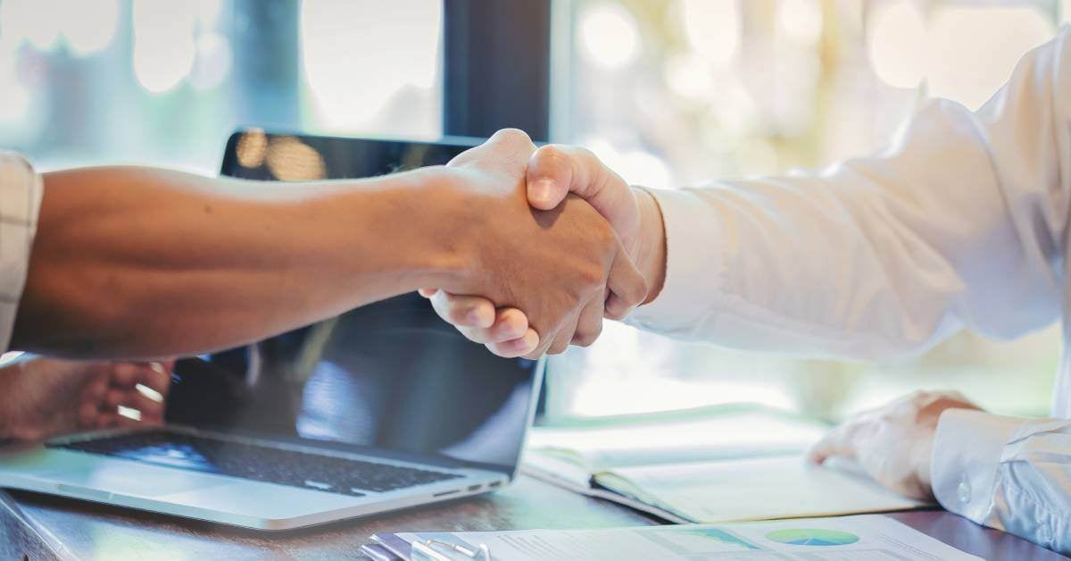 Two professionals share a firm handshake over a desk with a clipboard and laptop. They have come to a deal.