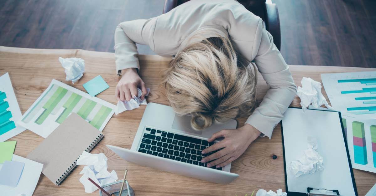 Top down of a woman with her head on her desk. Also on the desk is a laptop, forms, and several crumpled pieces of paper.