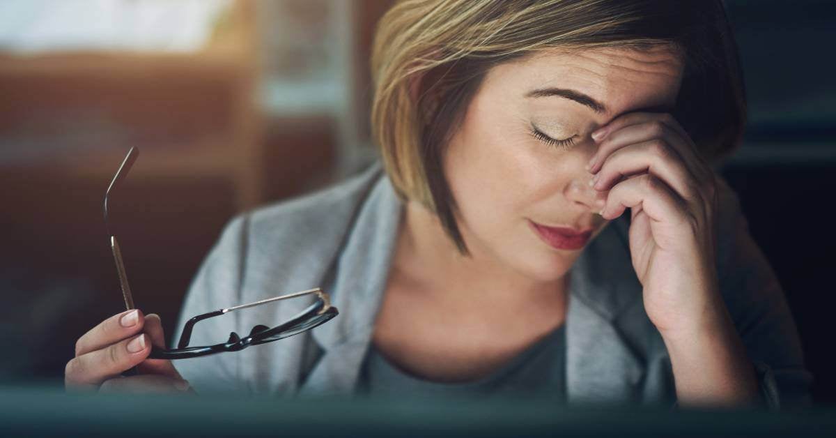 Woman at a computer. She's looking away from the computer and holding her hand to the bridge of her nose with her eyes closed.