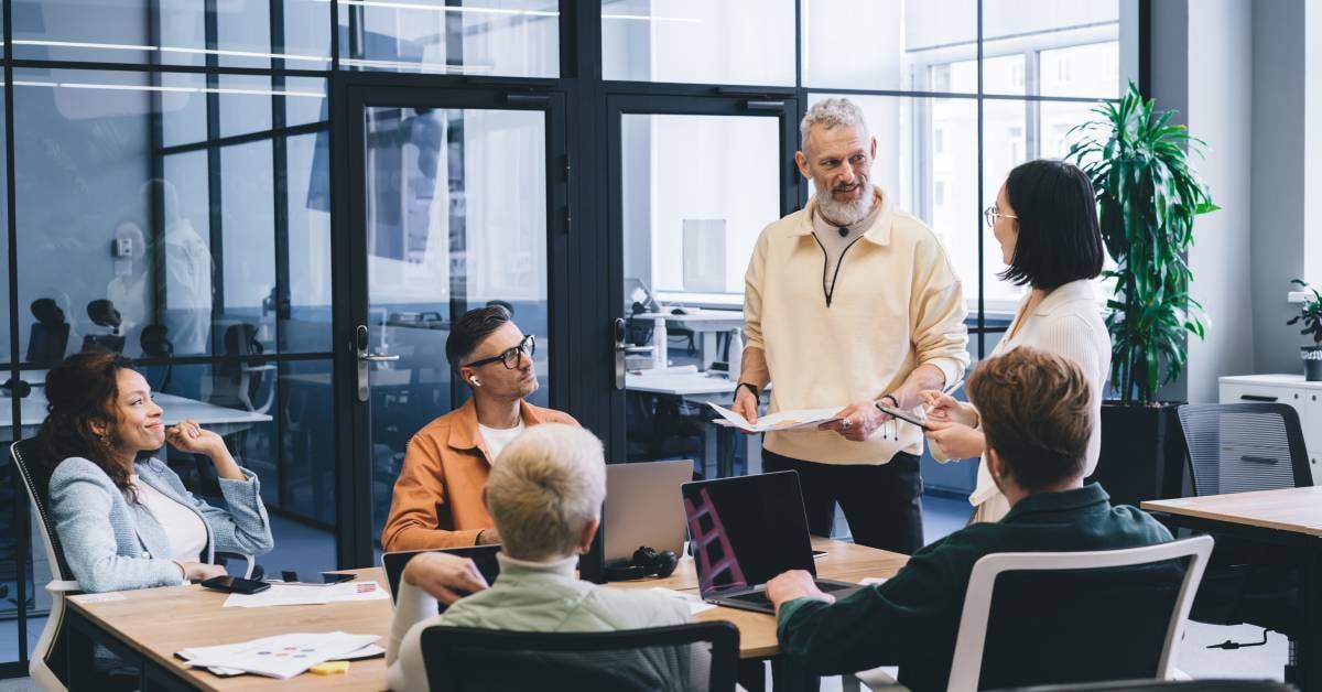 People sit at an office conference table. Two people are standing and looking at each other while holding papers.