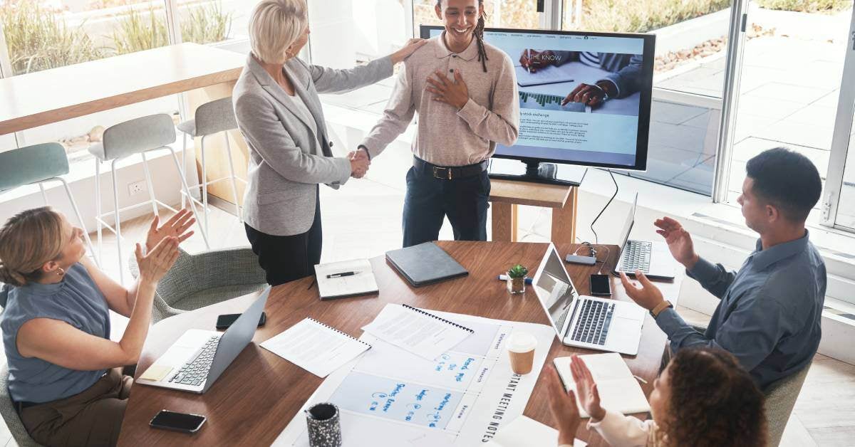 A group of employees are at a conference table. A woman is shaking a young person's hand and resting her hand on their shoulder.