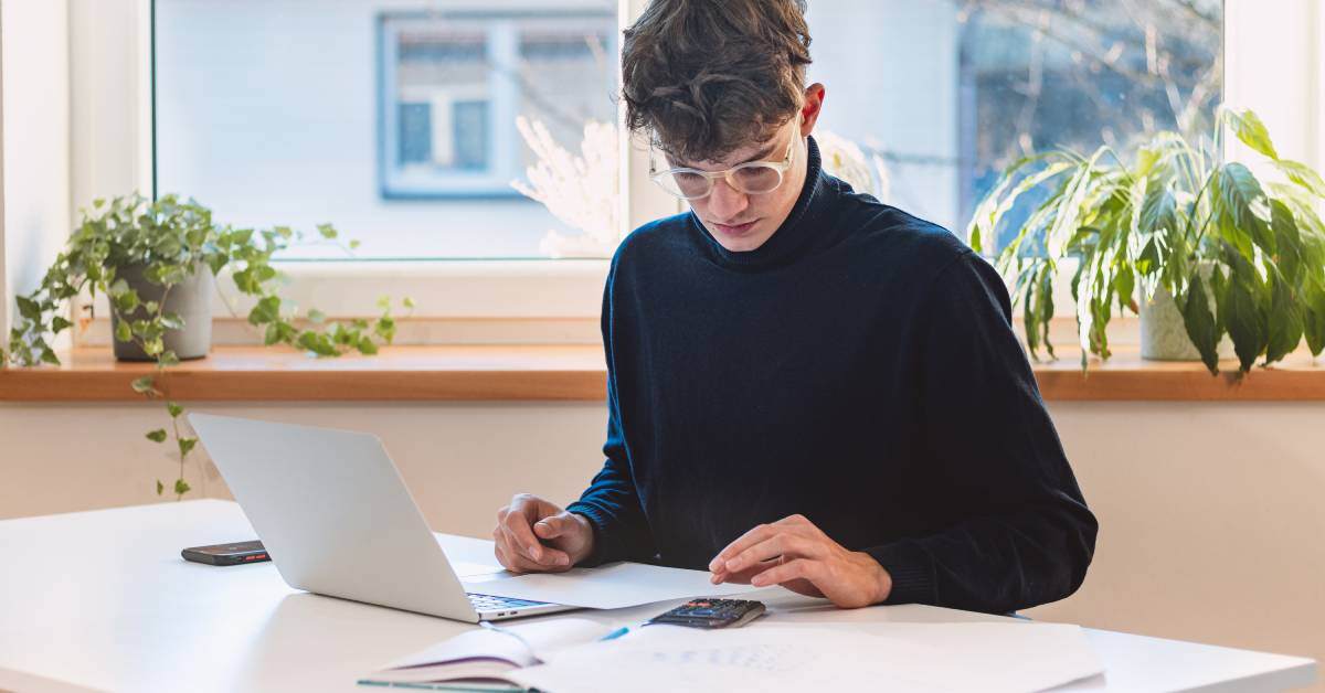 A young person in a black sweater sitting at a desk and typing on a calculator. The desk has a laptop and paperwork on it.