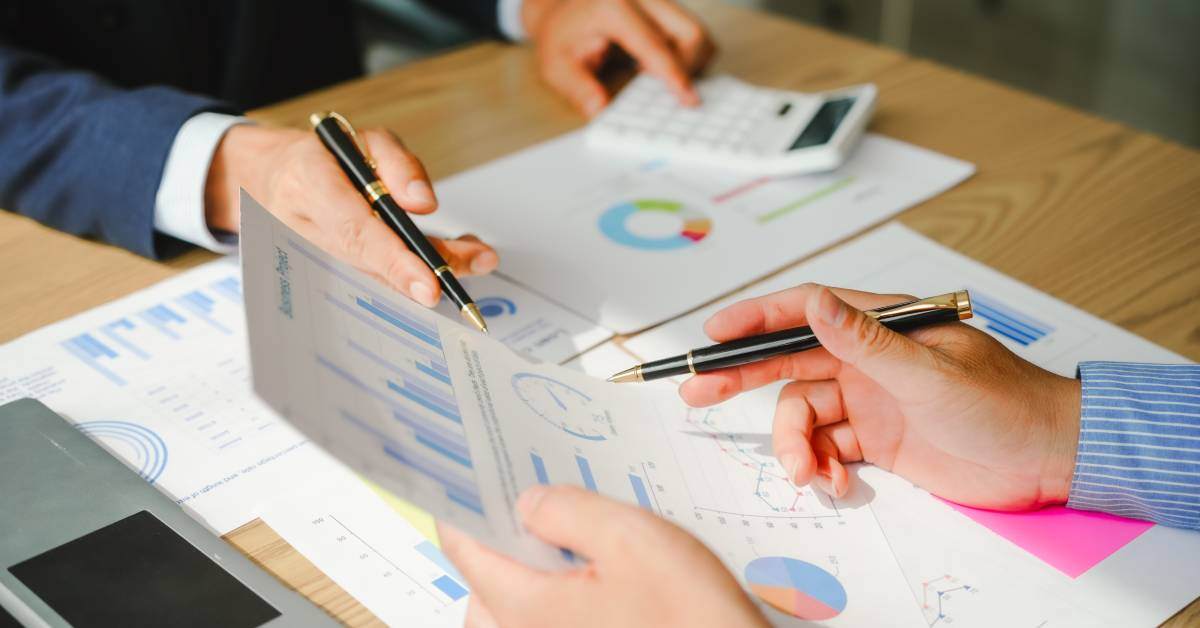 Business people sitting at a desk covered in forms with graphs on them. Two hands are using pens to point at a paper.