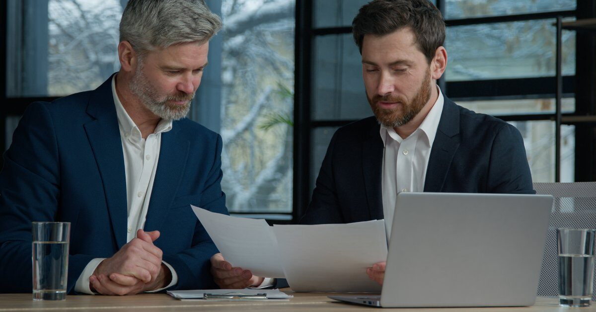 Two men in suits are sitting at a desk with a laptop on it. One is holding up some papers for them to look at.