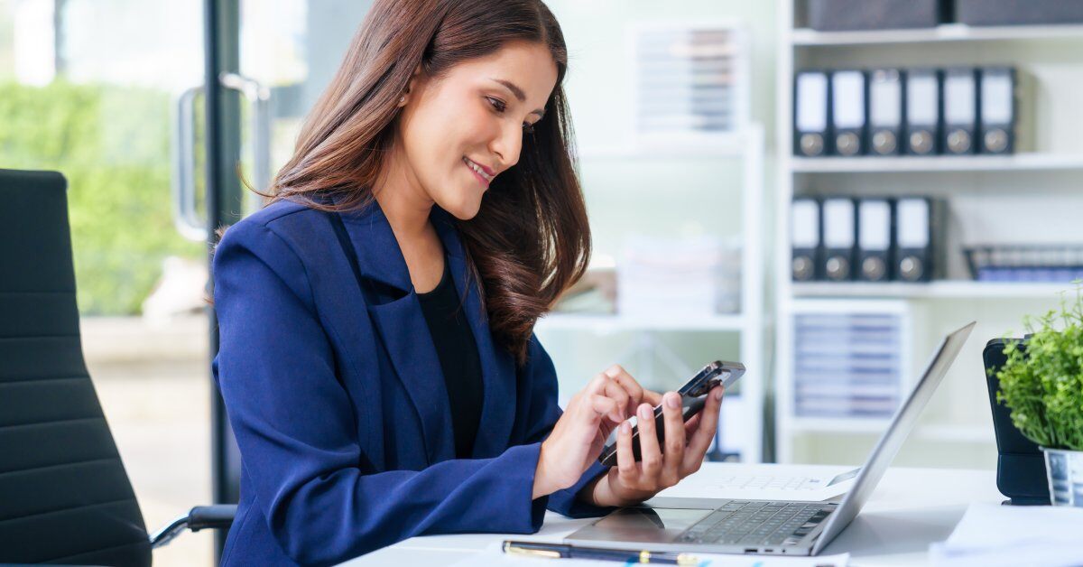 A young female executive in a bright blue blazer is sitting at a desk. She's using a smartphone next to an open laptop.