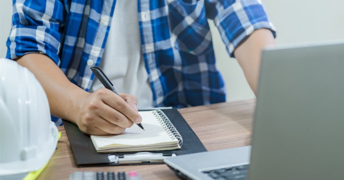 A man in a blue plaid shirt sits at a desk and looks at a laptop. With his right hand, he's writing in a notepad with a pen.