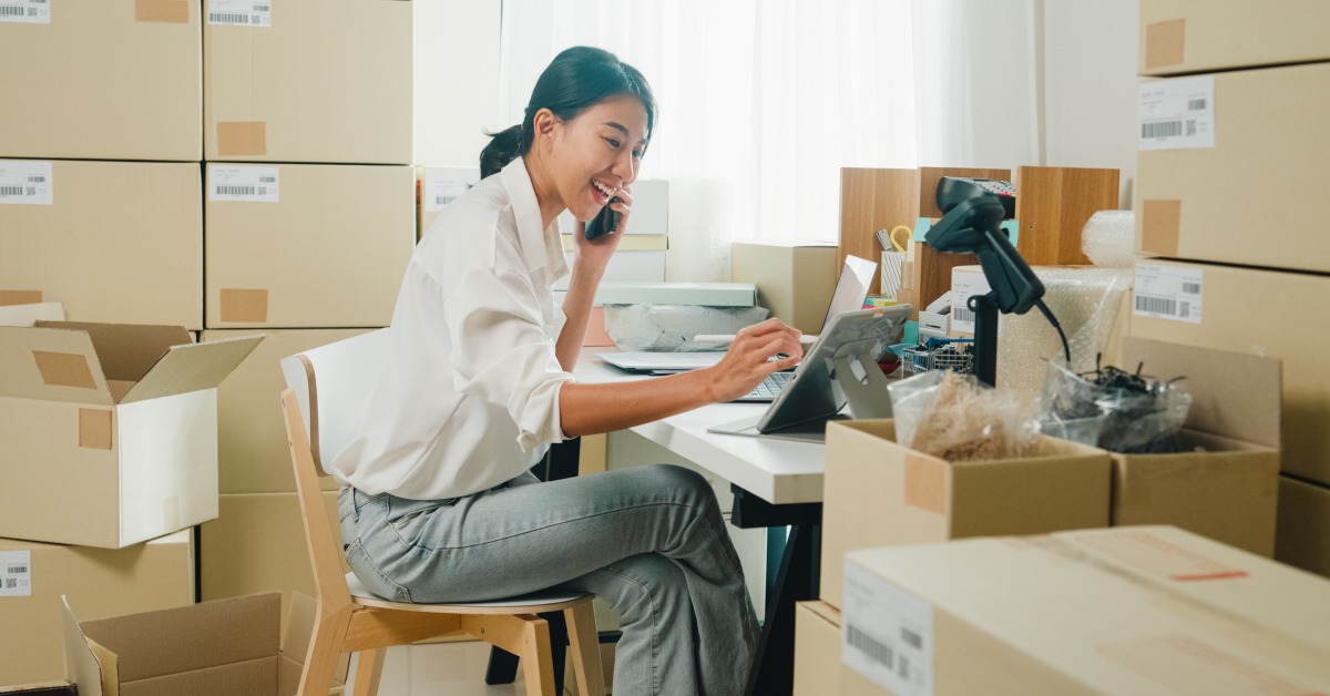 A woman using a phone and tablet at a desk surrounded by cardboard boxes, packing supplies, and window light.
