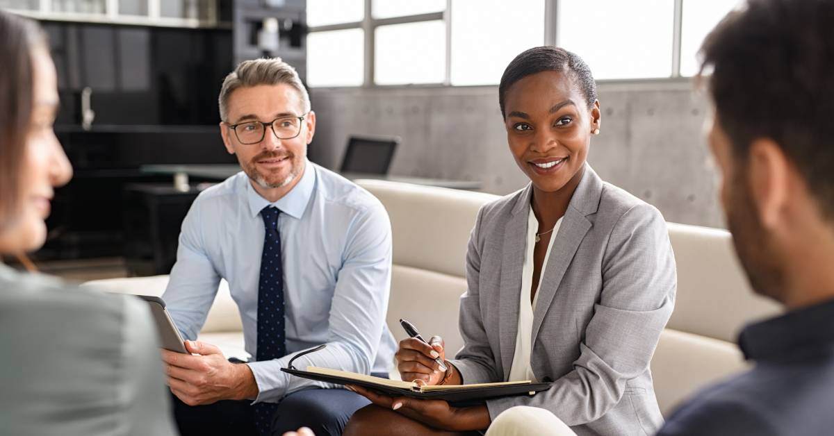 Four people in business dress holding paperwork smile, sit together, and discuss a potential business deal.