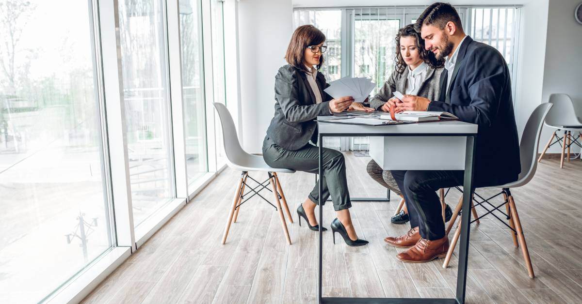 A pair of professionals are sitting at a desk in an open office and going over forms with a broker sitting across from them.