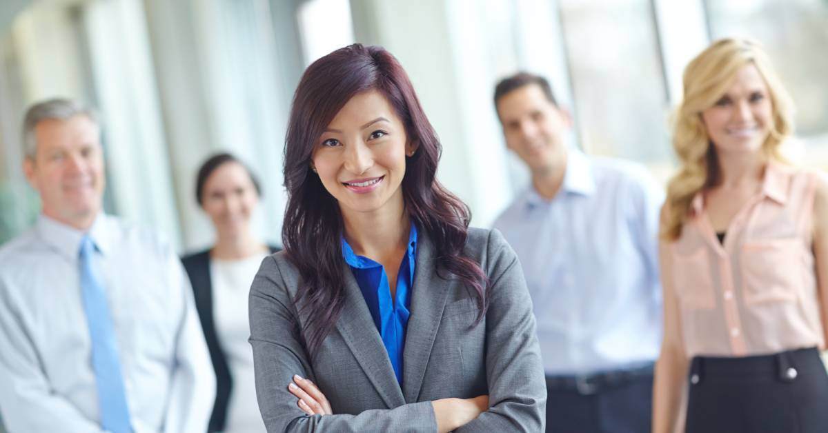 A woman in a professional blazer is standing with her arms crossed. Four other business people are standing, blurry, behind her.