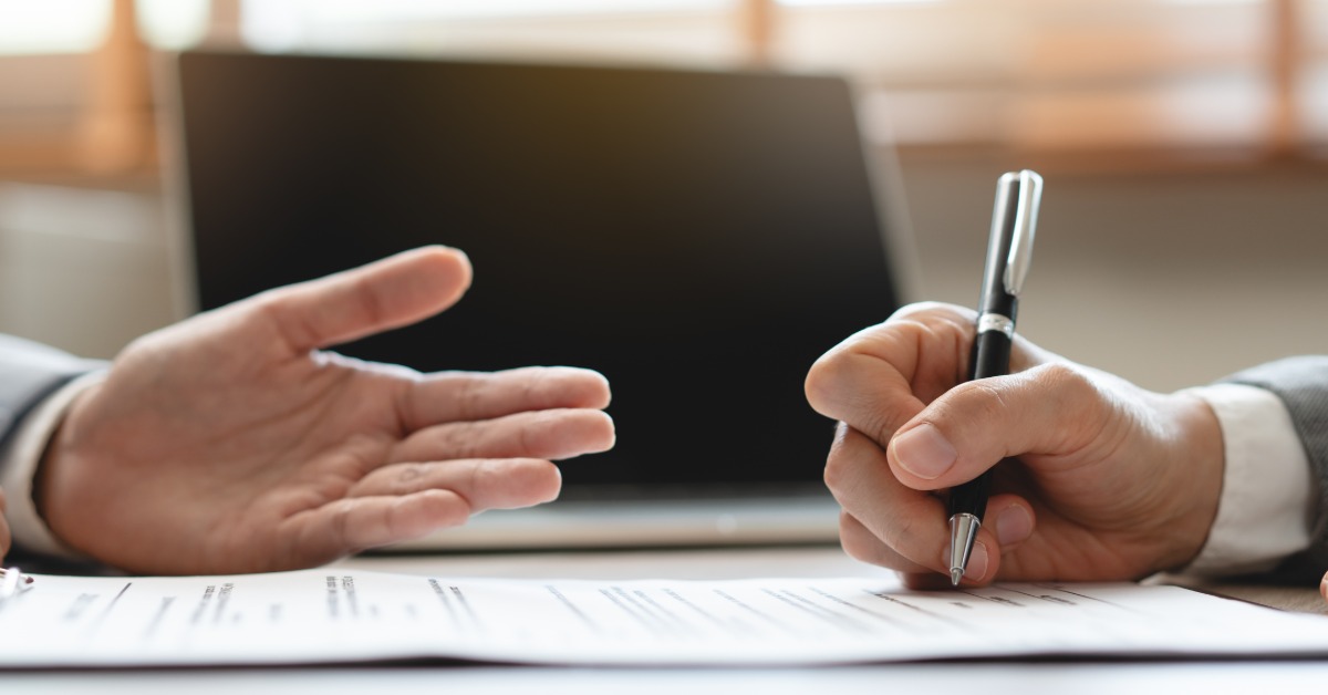 Two professionals sit at a table reviewing paperwork together as one person holds a pen near the bottom of the document.