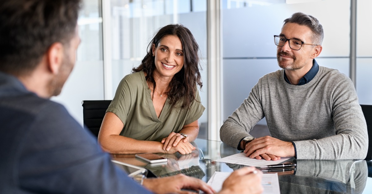 Three professionals sit around a glass conference table during a business meeting in a modern office setting.